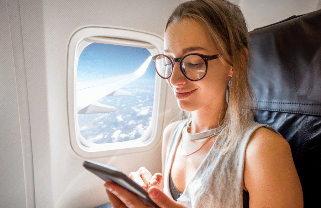 Young woman wearing eyeglasses who is sitting with phone on the airplane seat near the window during the flight - Budget Travel Tips