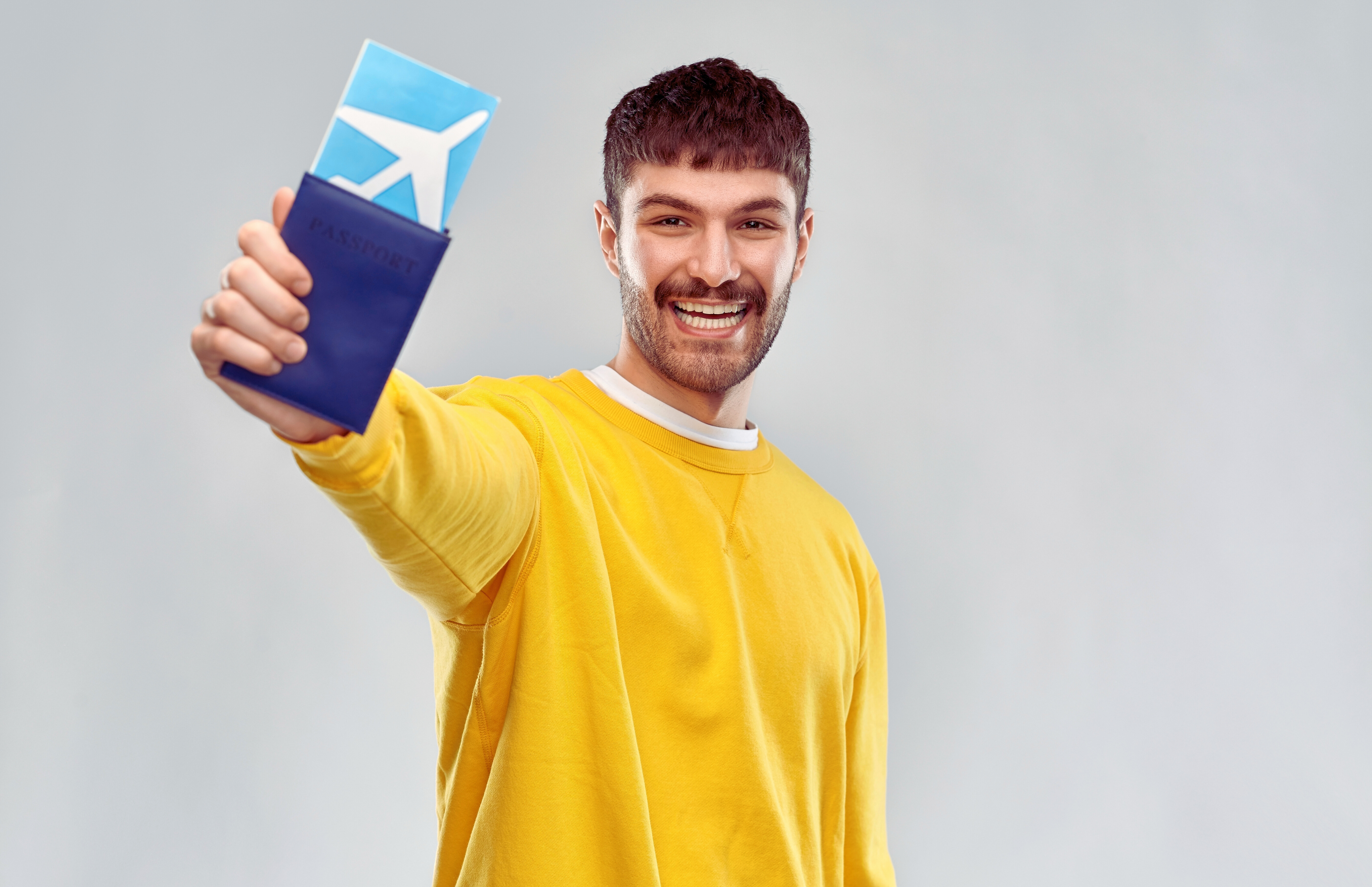 A young man smiling wearing a yellow sweatshirt holding an airline ticket and blue passport - Budget Travel Tips