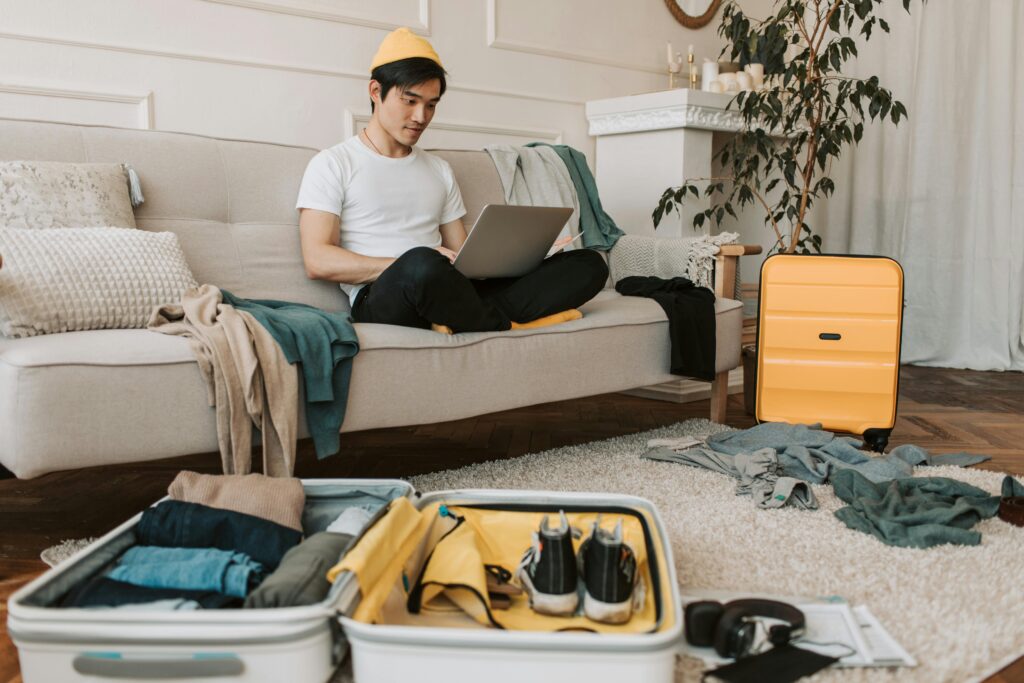House-Sitting and Home Exchange - A young man packing his luggage is sitting on a couch sending email on his laptop to communicate with the house owner
