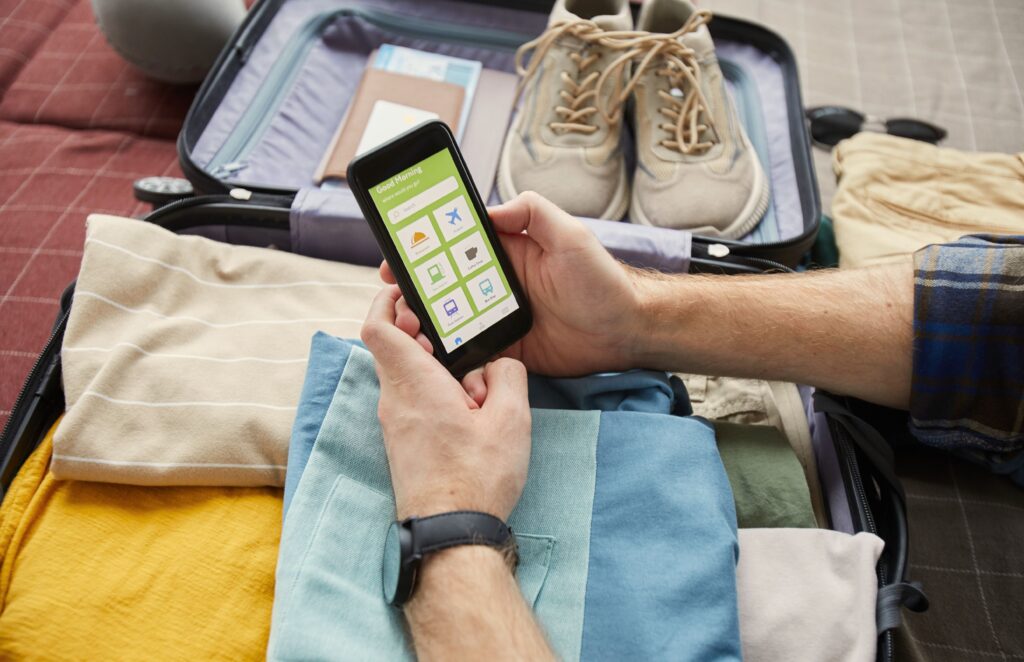 House-Sitting and Home Exchange - Hands of a man holding a mobile phone looking into an app while packing his travel essentials in the luggage