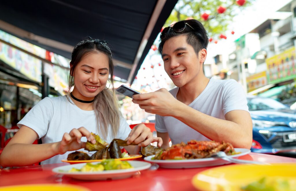 Cheap Travel Food Tips - Local Flavors - a young woman eating local street food while a young man is taking a photo on the food using his mobile phone