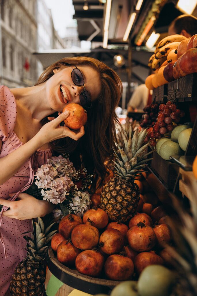 Food Travel Hacks - A young woman traveler smiling while holding a fruit and flowers in a local supermarket