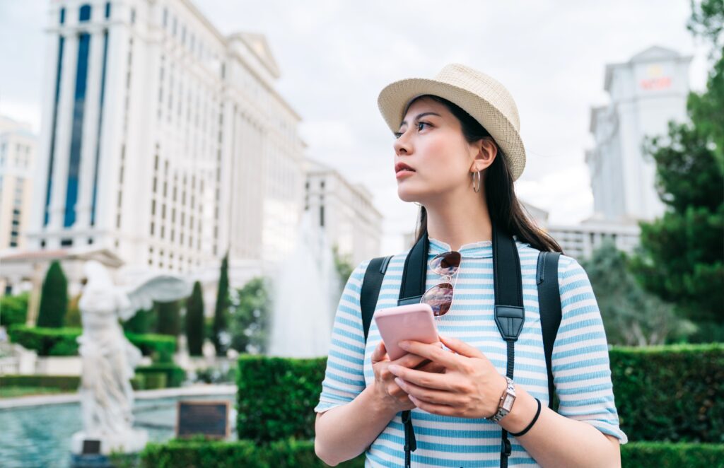 Free Attractions and Activities - Online Resources Apps - Pretty young woman traveler wearing a straw hat and holding a smart phone in front of fountain of a famous hotel. She is using her phone to search for direction.