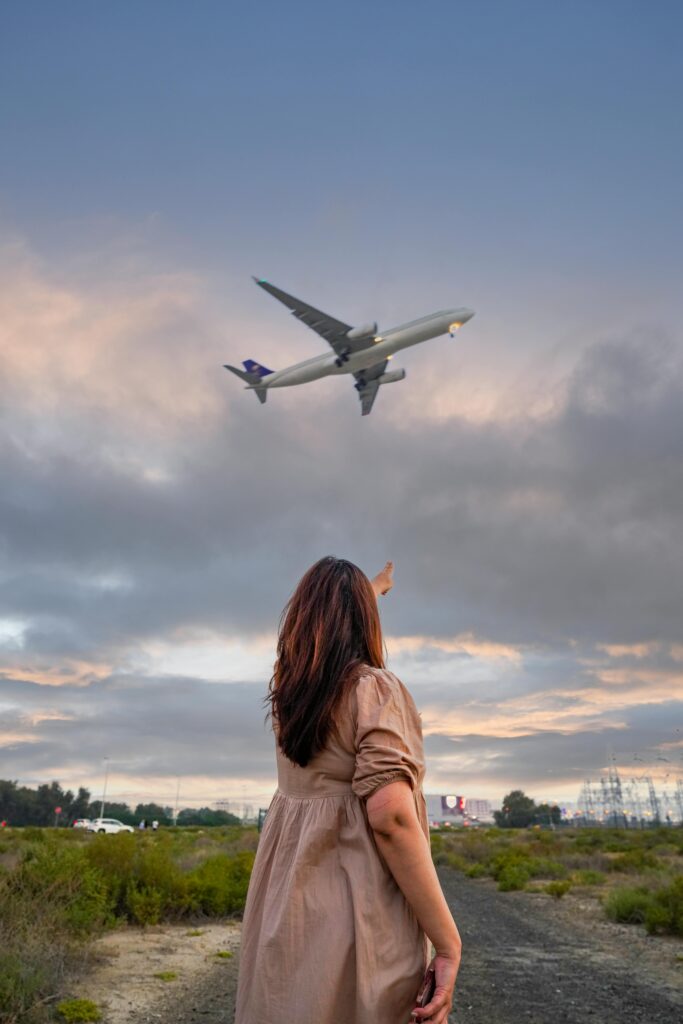 Track Cheap Flights - a young woman standing in an open field while pointing at an airplane flying in the sky