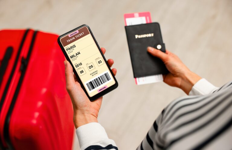 Traveletty - Best Free Travel Apps - Featured Image - Hands of a traveler holding a phone on the left hand, looking on a travel app while holding a passport on the right hand with a red luggage in the background.