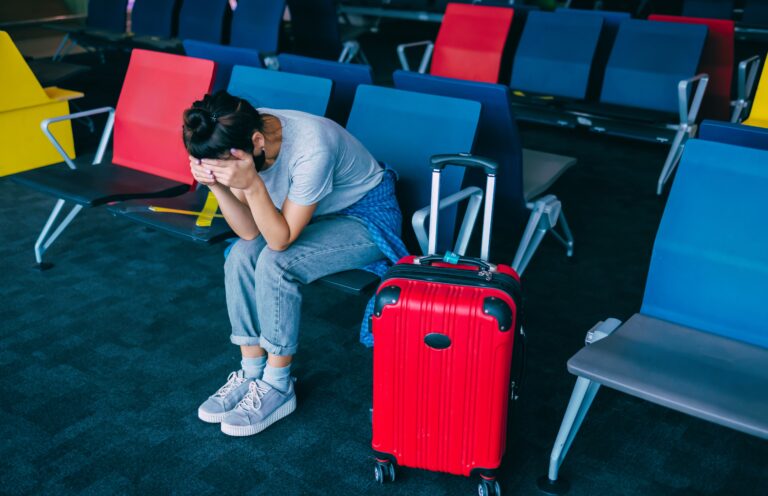 Traveletty - Common Travel Scams - Featured Image - A sad female tourist sitting that has red luggage beside her, being alone in the airport