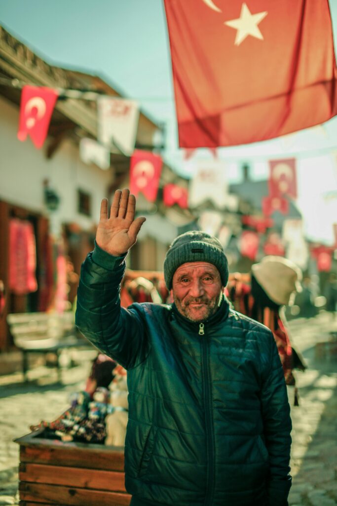 Traveletty - Travel Etiquette and Safety - an elderly man wearing a black jacket holding out his right arm to wave and greet while standing on the town street in Turkey