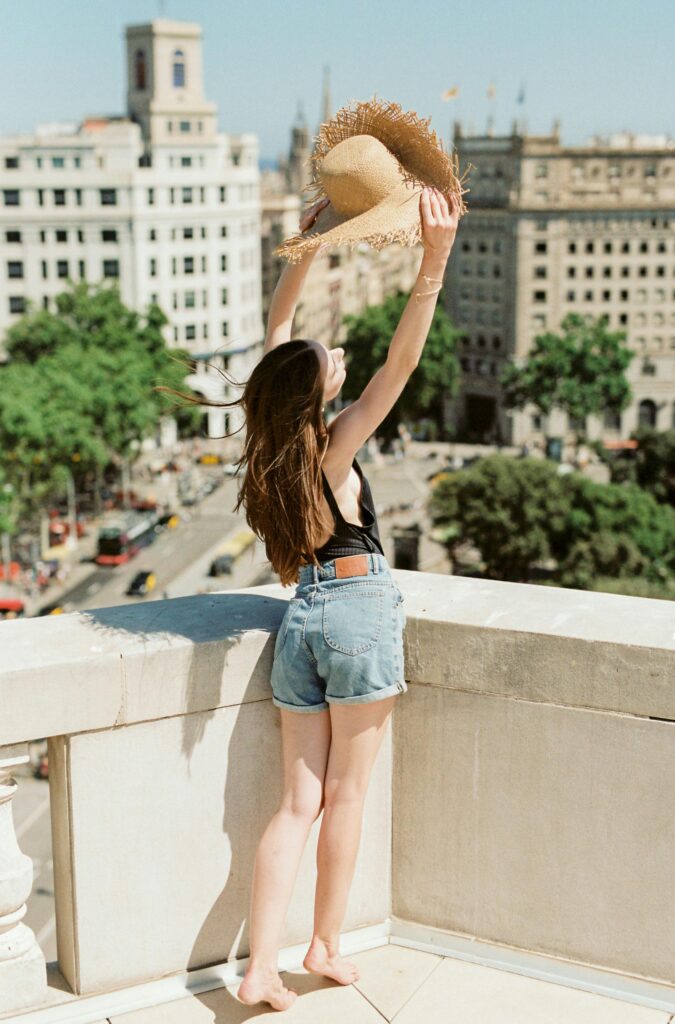 Traveletty - Travel Health and Safety - young female tourist in blue denim shorts and black tank top standing on rooftop holding up a hat