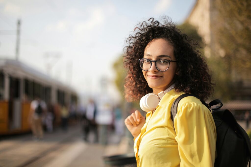 Traveletty - Budget Travel Transportation - A young smiling female traveler with curly hair wearing a yellow shirt standing at public transport terminal