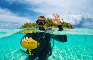 Traveletty - Dive Resorts - Destinations - scuba diver posing in the water while holding a coconut with Maldives engraved on it