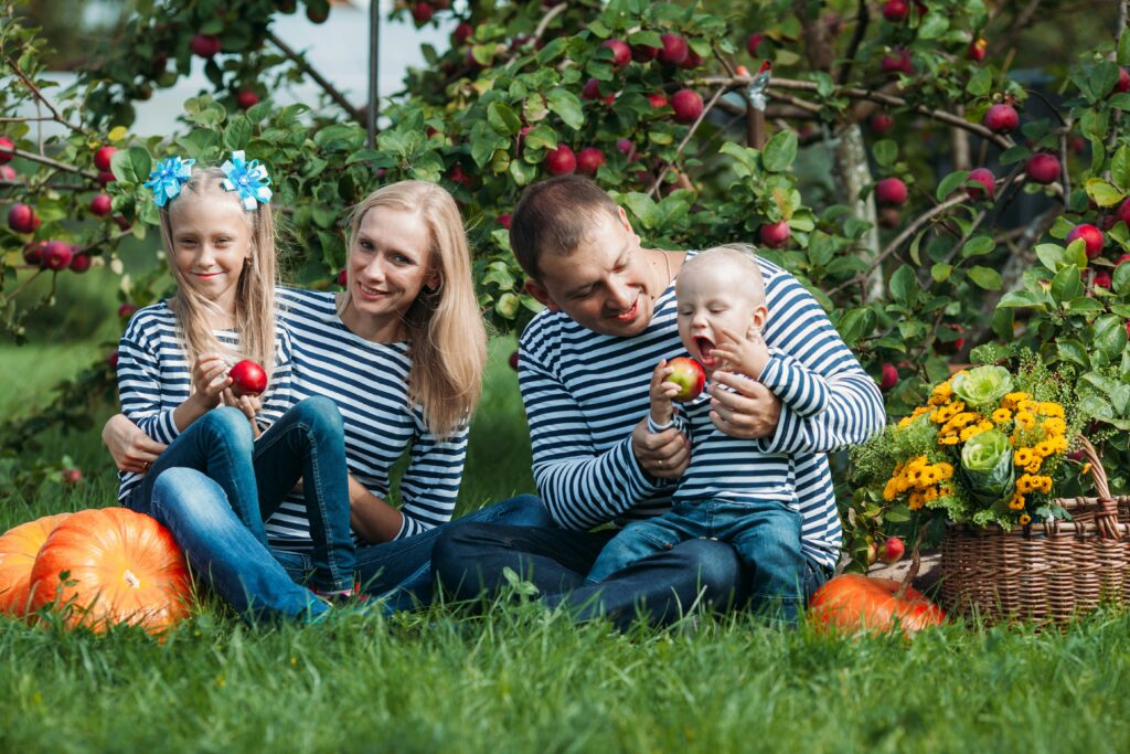 Traveletty - Farm Stays - Children posing with their parents, sitting on the grass surrounded by fruit bearing plants, vegetables and a basket of flowers.