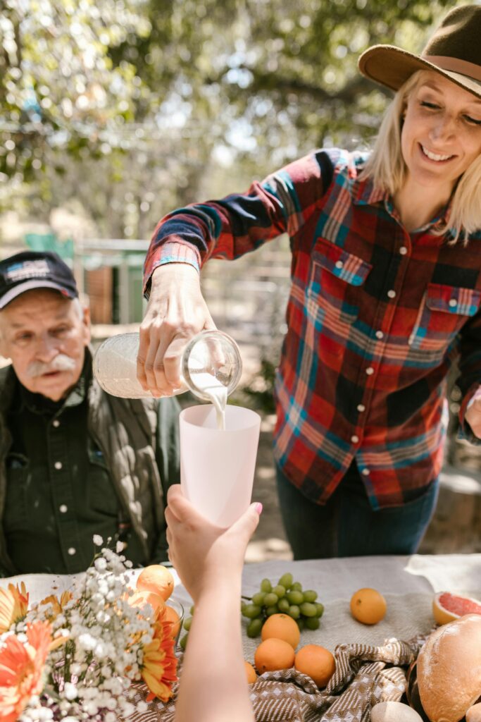 Traveletty - Farm Stays - woman pouring milk from a glass jar into a cup, surrounded by people gathering around a dining table with fruits, bread, and eggs on top to enjoy an outdoor meal in a farm stay